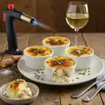 Individual ramekins of crab brûlée, topped with caramelized cheese and garnished with fresh chives, served on a white plate with a glass of white wine.